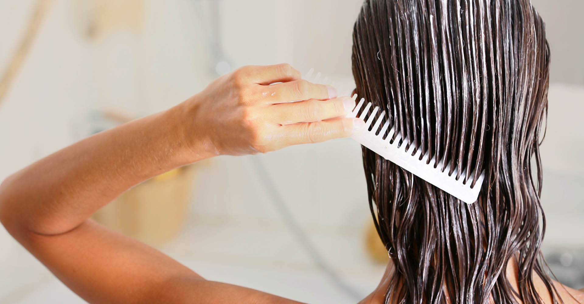Ragazza mora, vista di spalle, in doccia, con i capelli bagnati e il fluido applicato. La ragazza di sta pettinando i capelli